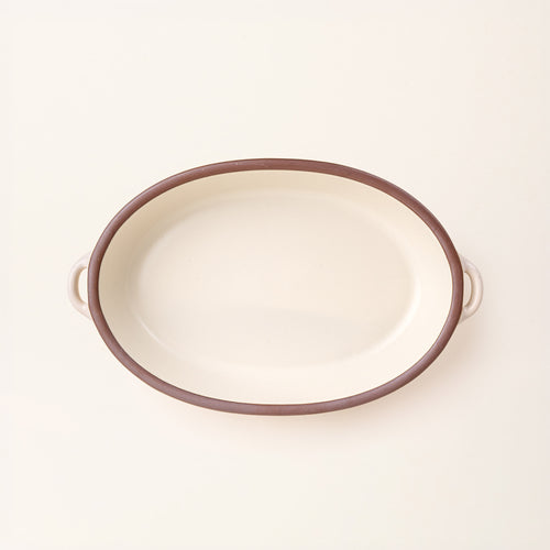 A large, oval, matte, warm off-white ceramic baking dish with two side handles, on a clean, solid off-white background.
