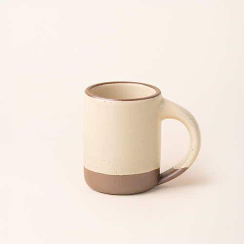 A medium sized ceramic mug with handle in a cream color featuring iron speckles and unglazed rim and bottom base.