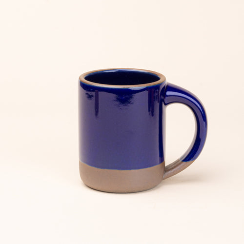 A big sized ceramic mug with handle in a glossy dark blue color featuring iron speckles and unglazed rim and bottom base.