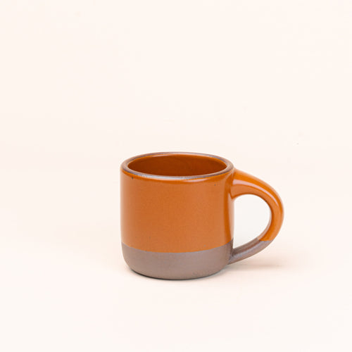 A small sized ceramic mug with handle in an earthy orange glaze featuring iron speckles and unglazed rim and bottom base.