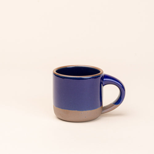 A small sized ceramic mug with handle in a glossy dark blue glaze featuring iron speckles and unglazed rim and bottom base.