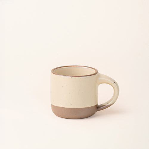 A small sized ceramic mug with handle in a cream color featuring iron speckles and unglazed rim and bottom base.
