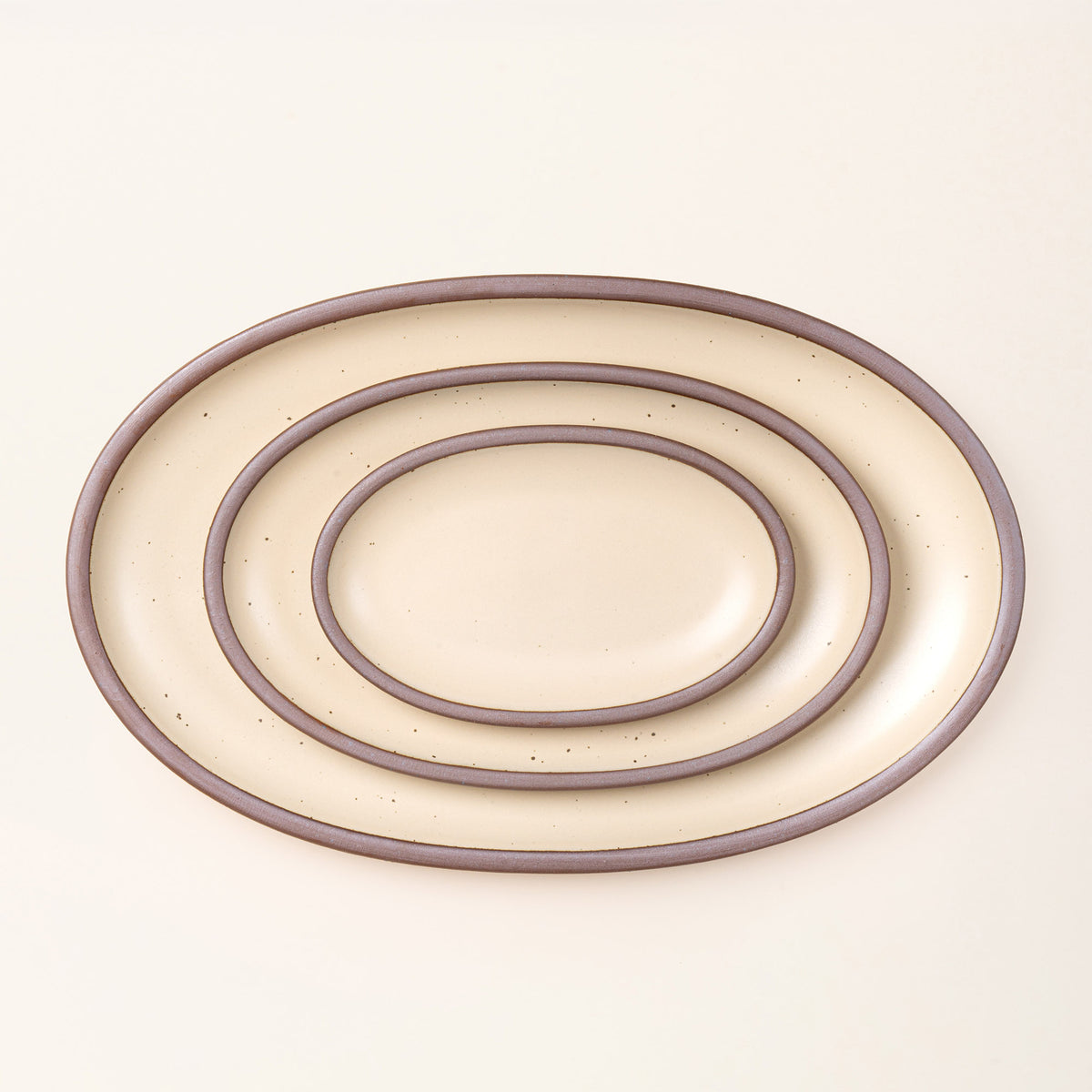 A large, medium, and small oval ceramic platter nesting in a cream color featuring iron speckles and an unglazed rim
