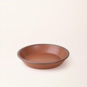 A shallow round ceramic pie plate in a matte terracotta color featuring an unglazed rim, photographed against a cream background.
