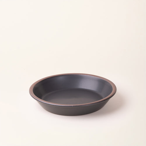 A shallow round ceramic pie plate in a matte graphite black color featuring an unglazed rim, photographed against a cream background.