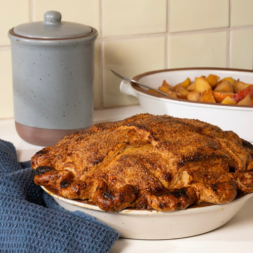 A baked fruit pie with a golden-brown crust sits in a warm off-white ceramic pie dish on a kitchen counter, with a blue cloth napkin, a speckled blue-gray lidded jar, and a bowl of fruit chunks in the background.