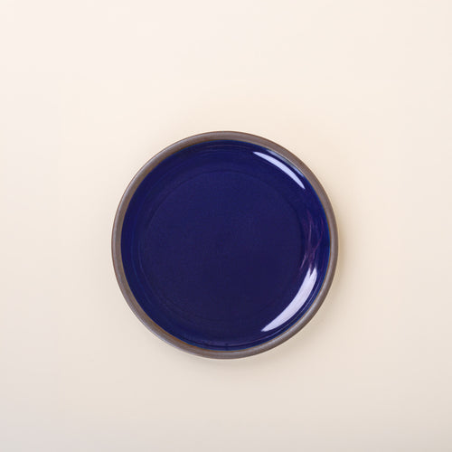 A medium sized ceramic plate in a glossy dark blue color featuring iron speckles and an unglazed rim.