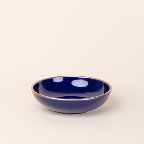 A dinner-sized shallow ceramic bowl in a glossy dark blue color featuring iron speckles and an unglazed rim