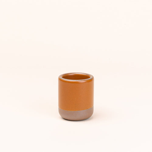 A small, short ceramic mug cup in an earthy orange color featuring iron speckles and unglazed rim and bottom base.