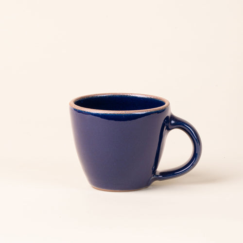 Round stoneware mug with handle, in an glossy dark blue color with iron speckles and an unglazed rim.