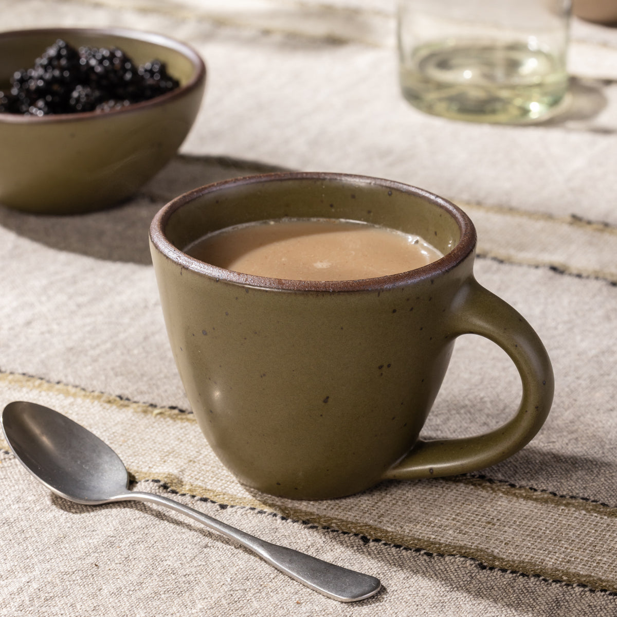 Olive-glazed stoneware mug with a rounded silhouette and speckled finish, styled with coffee on a rustic linen table.