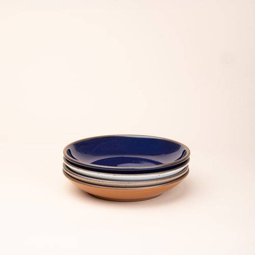 A stack of 4 medium ceramic plates with a curved bowl edge in an earthy orange, glossy dark blue, white, and warm pale brown colors featuring iron speckles and an unglazed rim.