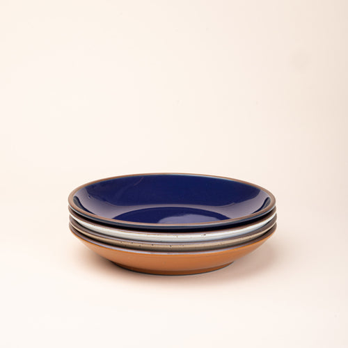 A stack of 4 large ceramic plates with a curved bowl edge in an earthy orange, glossy dark blue, white, and warm pale brown colors featuring iron speckles and an unglazed rim.
