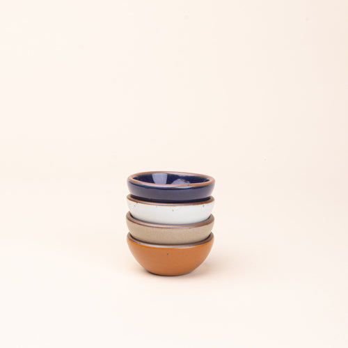 A stack of 4 tiny rounded ceramic bowls in an earthy orange, glossy dark blue, white, and warm pale brown colors featuring iron speckles and an unglazed rim