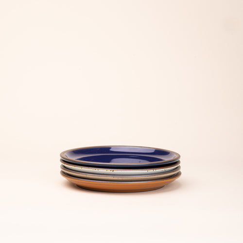 4 medium sized ceramic plates in a glossy dark blue, earthy orange, warm pale brown and white colors featuring iron speckles and an unglazed rim