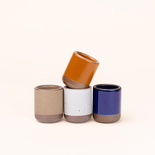 A set of 4 small, short ceramic mug cups in earthy orange, glossy dark blue, white, and warm pale brown colors featuring iron speckles and unglazed rim