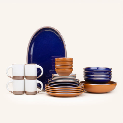 A 26-piece tablesetting with four mugs, four cake plates, four side plates, four dinner plates, 4 shallow bowls, 4 ice cream bowls, a large shallow bowl and an oval platter in a white, earthy orange, and glossy dark blue colors.
