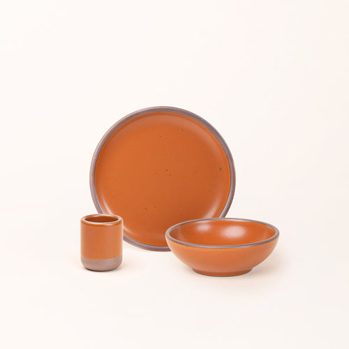 A small tiny cup, a medium sized plate, and a small breakfast bowl paired together in an earthy orange color featuring iron speckles.