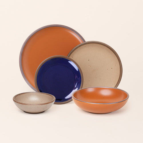 A breakfast bowl, everyday bowl, cake plate, side plate and dinner plate paired together in earthy orange, pale warm brown and glossy dark blue colors featuring iron speckles
