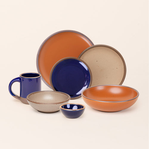 A breakfast bowl, everyday bowl, cake plate, side plate, dinner plate, mug and bitty bowl, paired together in earthy orange, pale warm brown and glossy dark blue colors featuring iron speckles.