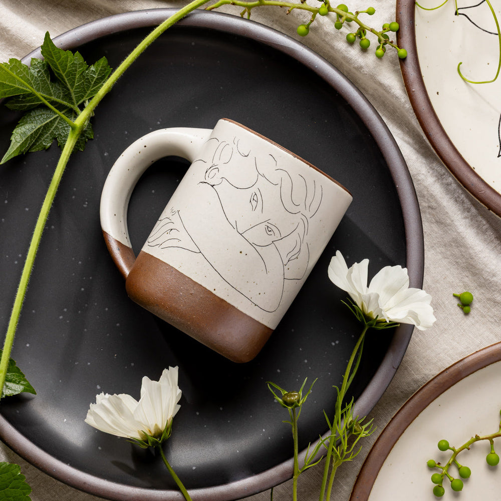 East Fork’s The Mug in a warm off white color with handle with a black decal of Henri Matisse’s Jeune femme la visage enfoui dans les bras, a bright blue, a line drawing of a young woman with face buried in her arms, on the body of the mug, styled on a black plate with flowers