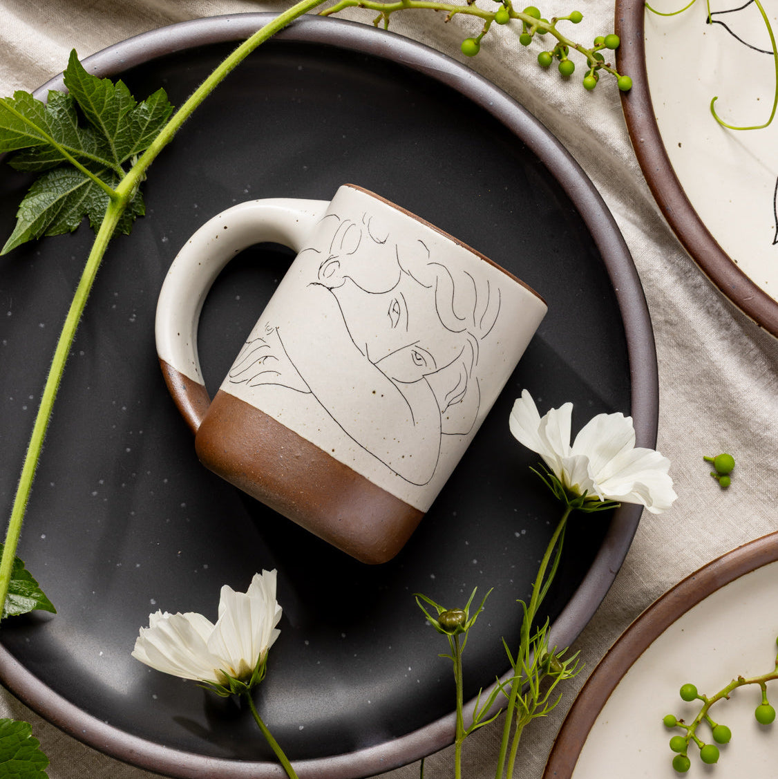 East Fork’s The Mug in a warm off white color with handle with a black decal of Henri Matisse’s Jeune femme la visage enfoui dans les bras, a bright blue, a line drawing of a young woman with face buried in her arms, on the body of the mug, styled on a black plate with flowers