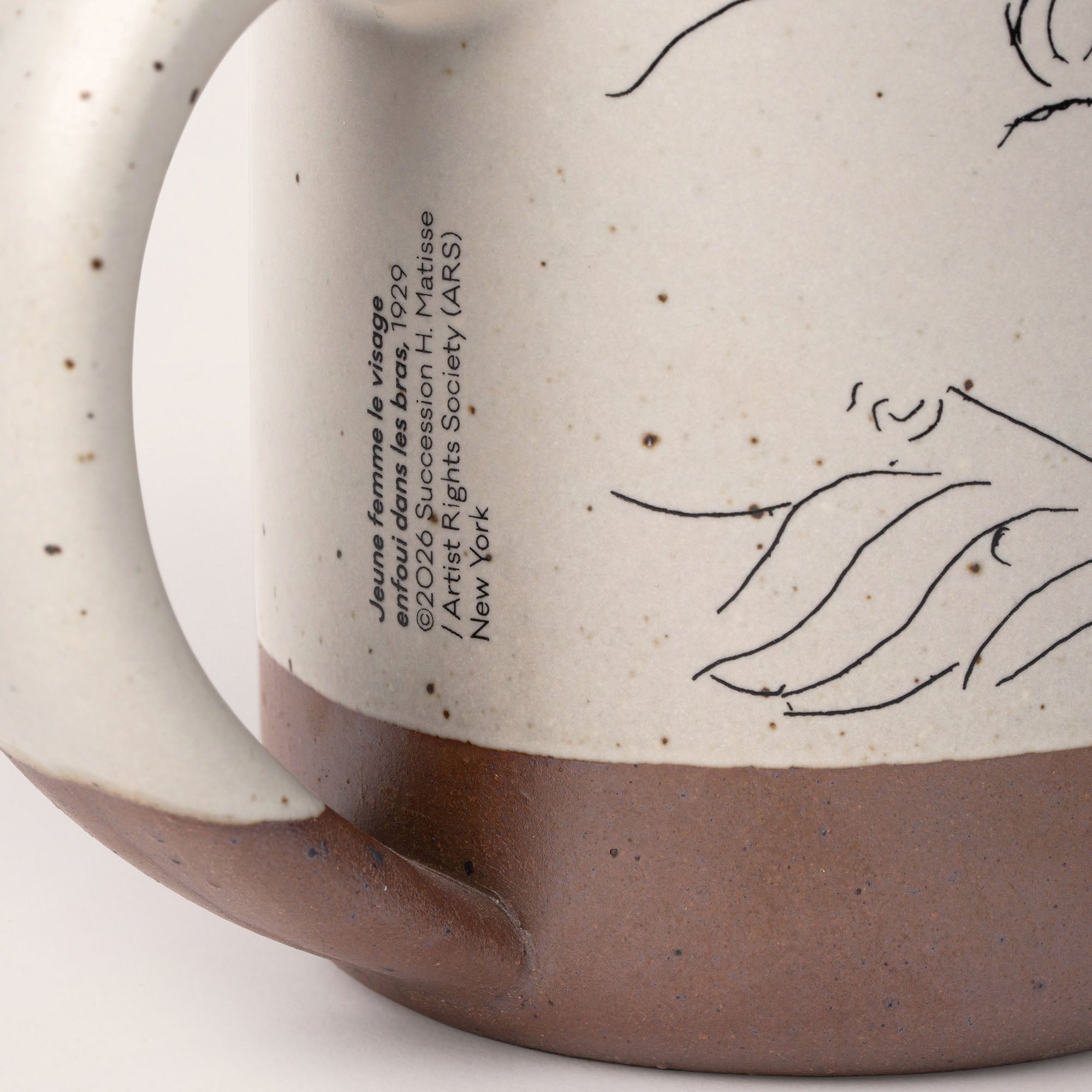 Closeup of the title and copyright under the handle of East Fork’s The Mug in a warm off white color with handle with a black decal of Henri Matisse’s Jeune femme la visage enfoui dans les bras, a bright blue, a line drawing of a young woman with face buried in her arms, on the body of the mug