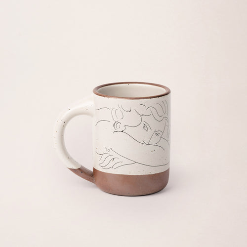 East Fork’s The Mug in a warm off white color with handle with a black decal of Henri Matisse’s Jeune femme la visage enfoui dans les bras, a bright blue, a line drawing of a young woman with face buried in her arms, on the body of the mug