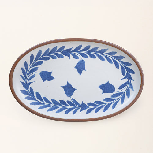 Oval ceramic platter with a hand-painted blue leaf and flower border on a white background with an exposed clay rim.