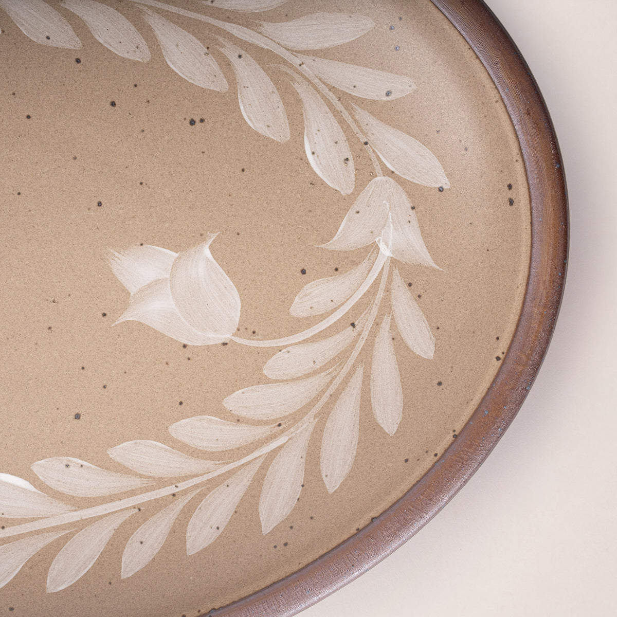 Close up of an Oval ceramic platter with a hand-painted white leaf and flower border on a warm pale brown background with an exposed clay rim.