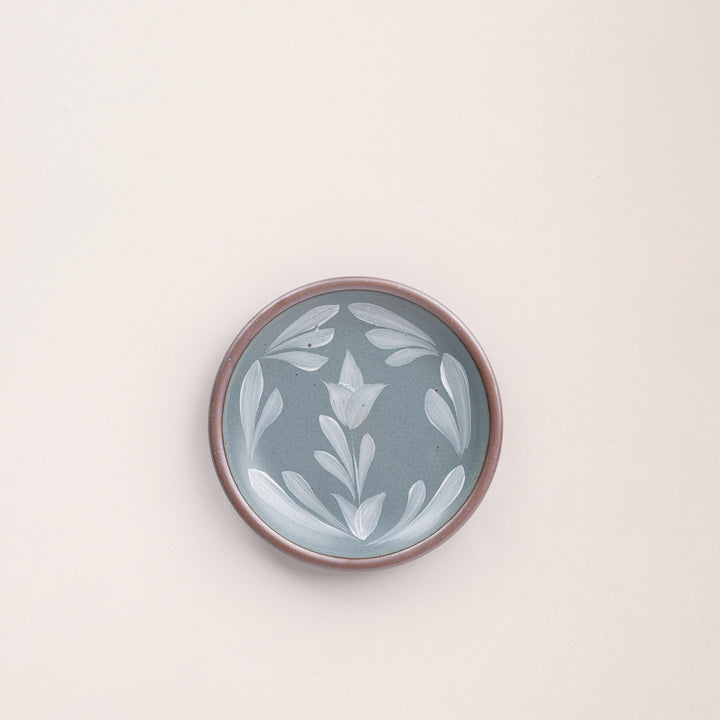 Small round ceramic plate with a hand-painted white flower and leaf design on a blue-grey background with an exposed clay rim.