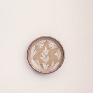 Small round ceramic plate with a hand-painted white flower and leaf design on a warm pale brown background with an exposed clay rim.