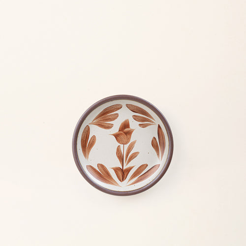 Small round ceramic plate with a hand-painted terracotta flower and leaf design on a cream background with an exposed clay rim.