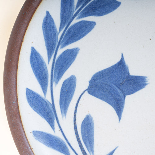 Closeup of a side round ceramic plate with a hand-painted blue leaf wreath and floral design on a white background with an exposed clay rim.