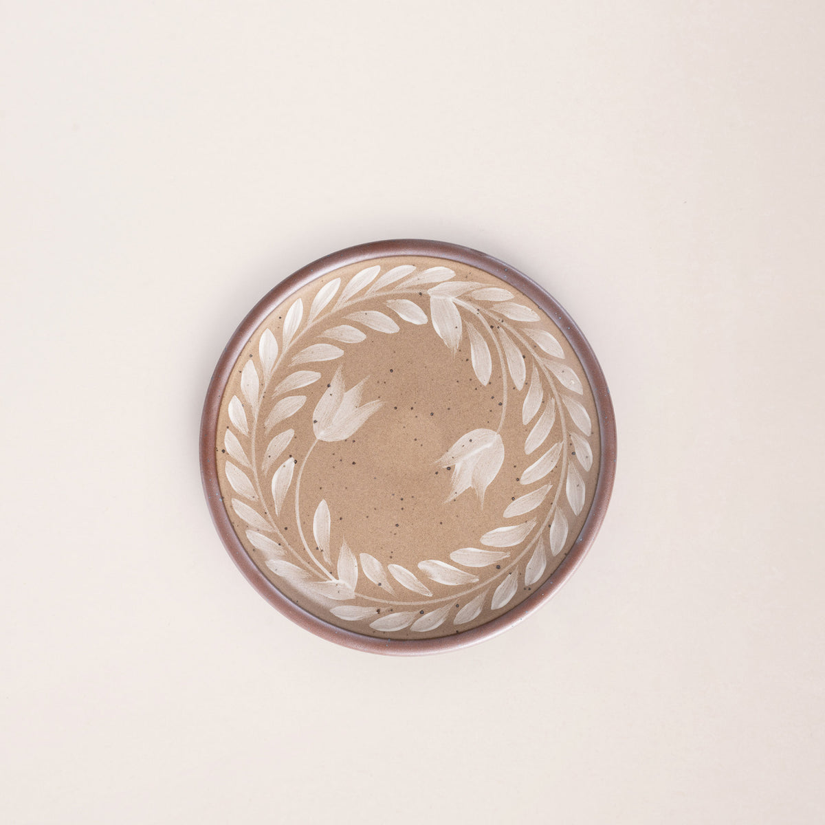 Side round ceramic plate with a hand-painted white leaf wreath and floral design on a warm pale brown background with an exposed clay rim.