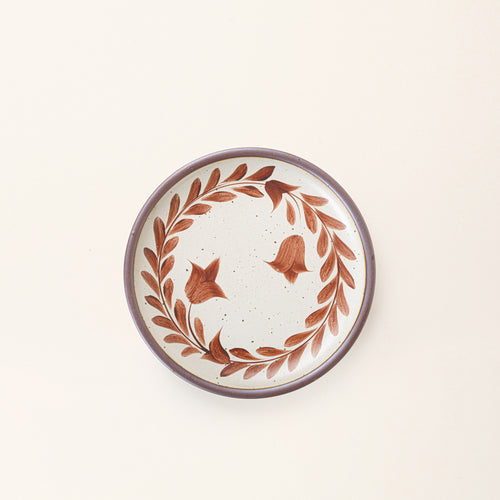 Side round ceramic plate with a hand-painted terracotta leaf wreath and floral design on a cream background with an exposed clay rim.