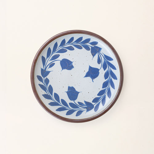 Dinner round ceramic plate with a hand-painted blue leaf wreath and floral design on a white background with an exposed clay rim.