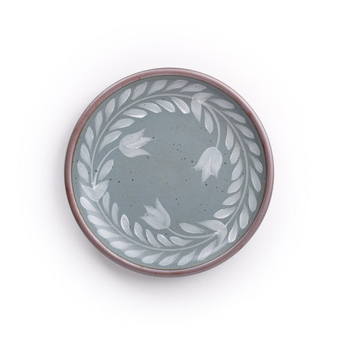 Dinner round ceramic plate with a hand-painted white leaf wreath and floral design on a blue-grey background with an exposed clay rim.