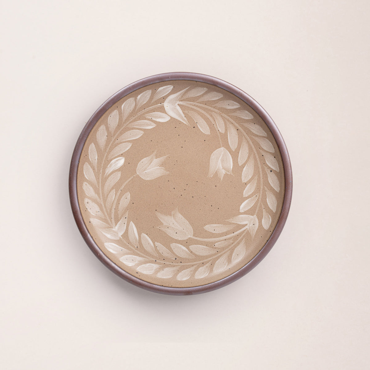 Dinner round ceramic plate with a hand-painted white leaf wreath and floral design on a warm pale brown background with an exposed clay rim.
