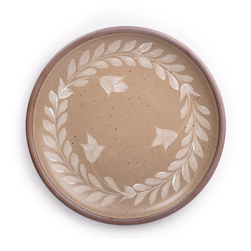 Large round ceramic platter with a hand-painted white leaf wreath and floral design on a warm pale brown background with an exposed clay rim.