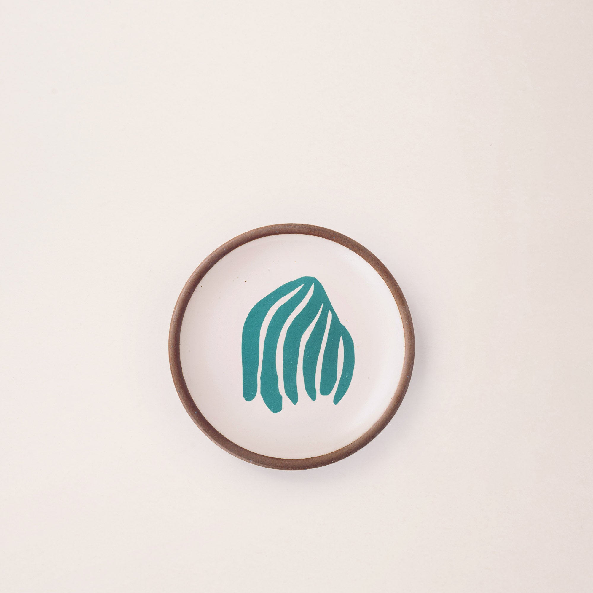 A ceramic dessert sized plate in a warm off-white color with unglazed rim, a teal frond-like cut-outs from Henri Matisse’s La Perruche et la sirène.