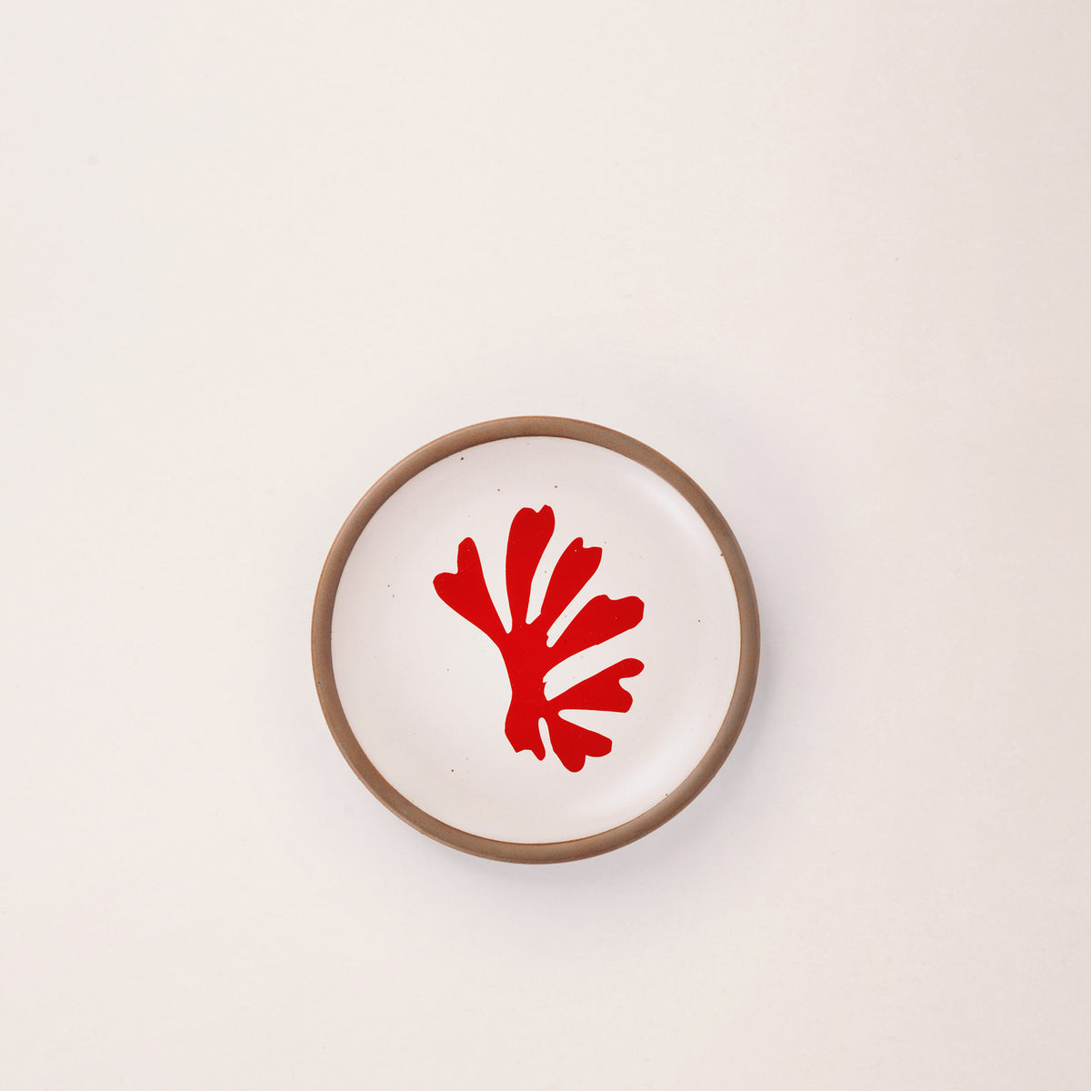 A ceramic dessert sized plate in a warm off-white color with unglazed rim, a red frond-like cut-outs from Henri Matisse’s La Perruche et la sirène.