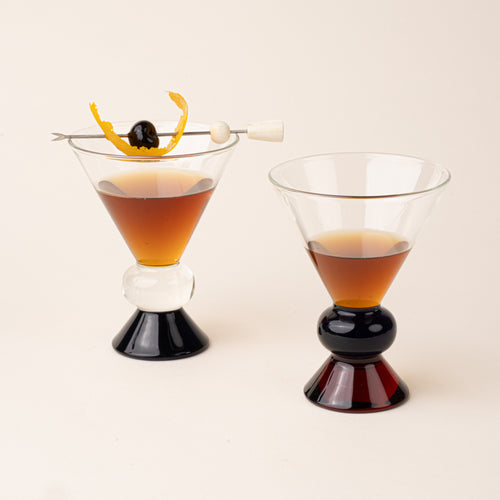 Two clear cocktail-style glasses with sculptural clear, blue and amber base. Glasses are filled with an amber drink and garnished with a curled orange peel and a cherry on a skewer.