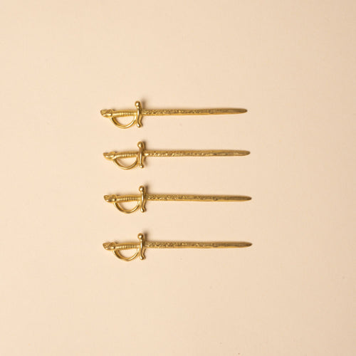 Set of four brass cocktail picks shaped like miniature swords, laid in a row on a neutral background.
