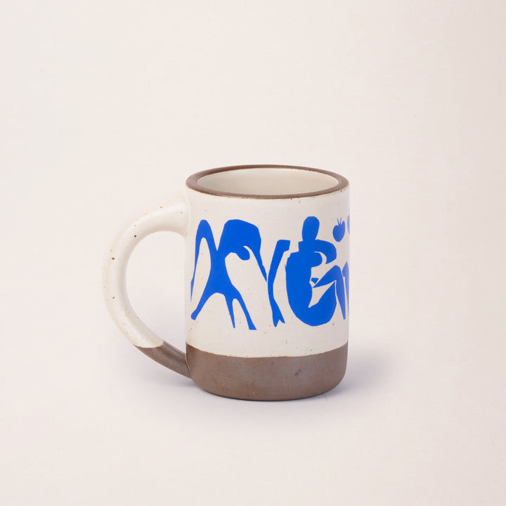 East Fork’s The Mug in a warm off white color with handle with a blue decal of Henri Matisse’s Femmes et singes, a bright blue, abstract cut-out figures of women sitting, monkeys, and pomegranate across the body of the mug
