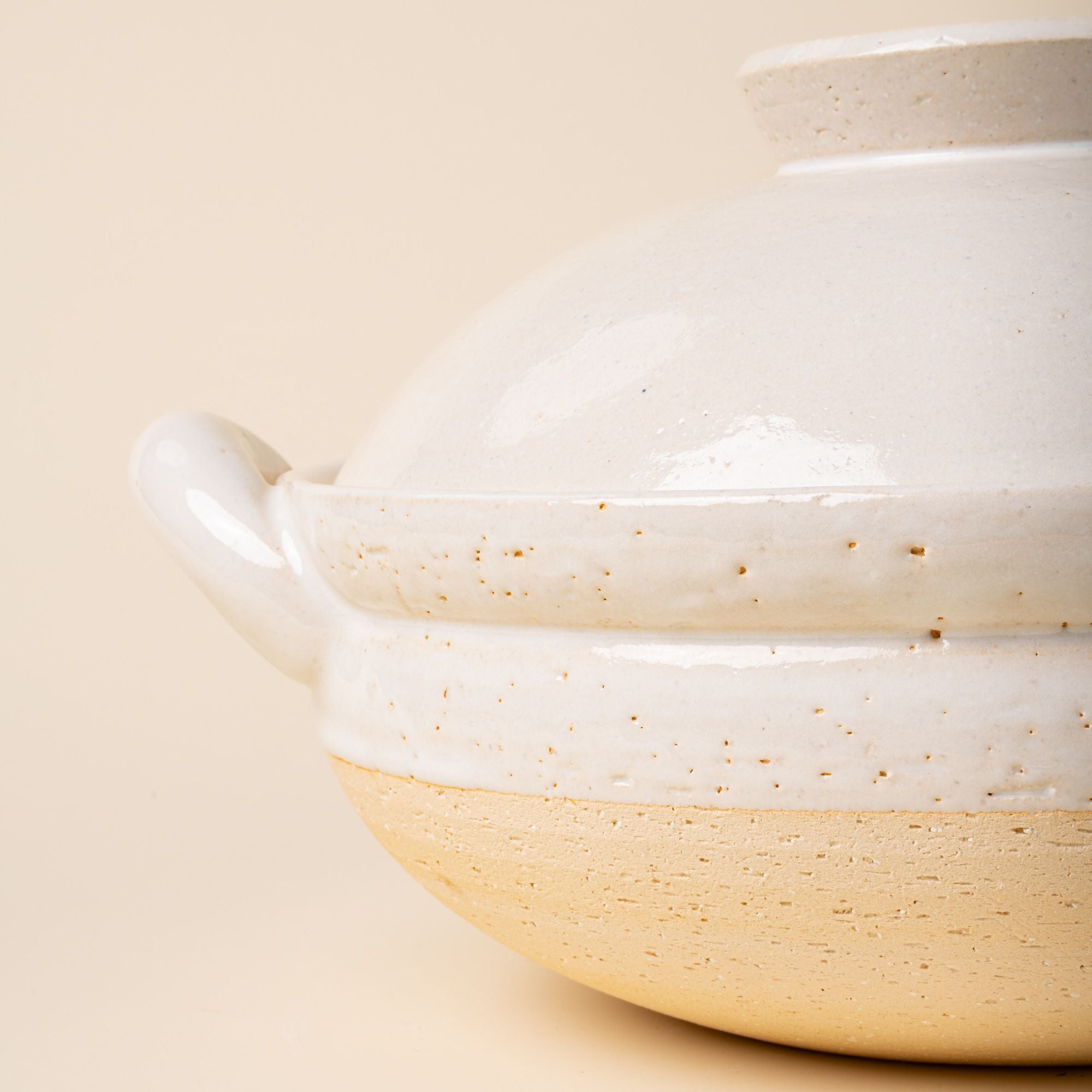 A closeup of the side of a white ceramic lidded donabe steamer with side handles