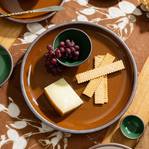 Glossy caramel brown ceramic platter with crackers, cheese, and grapes on a floral table runner.