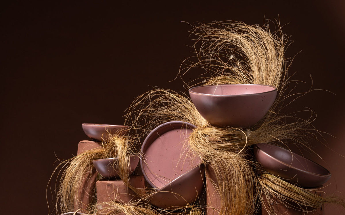 Stack of dusty mauve pink ceramic bowls and bowls piled together with strands of dried grass against a dark background.