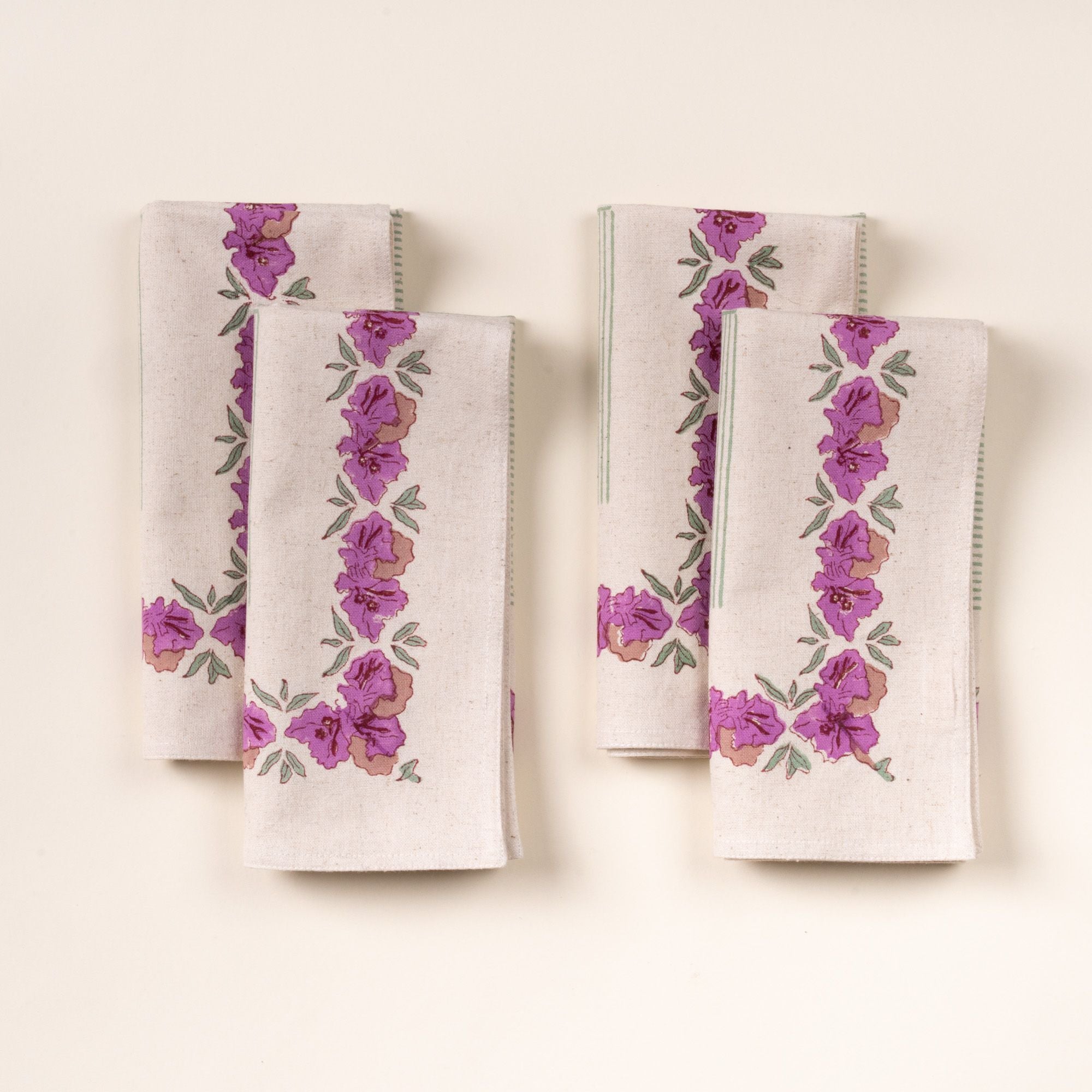 Four folded dinner napkins with purple floral borders and green stitched edges, arranged in two pairs on a cream background.