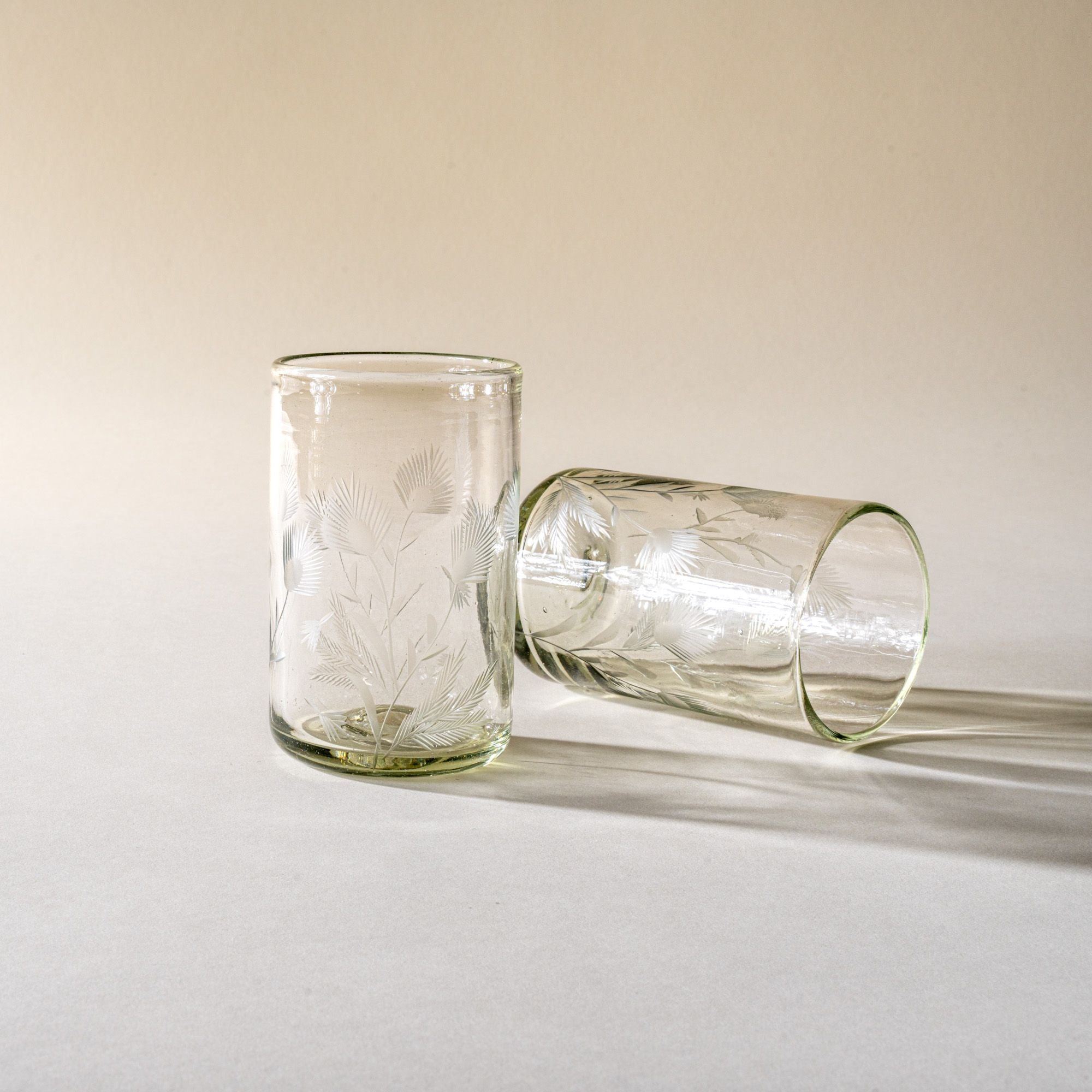 Two clear tumbler glasses made of recycled glass with etched thistle designs on it, one standing up, one laying on the side.